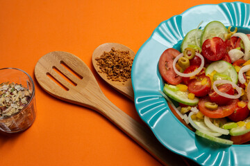 Um prato com salada de vegetais, um vidro com temperos desidratados e dois talheres de madeira sobre um fundo alaranjado.