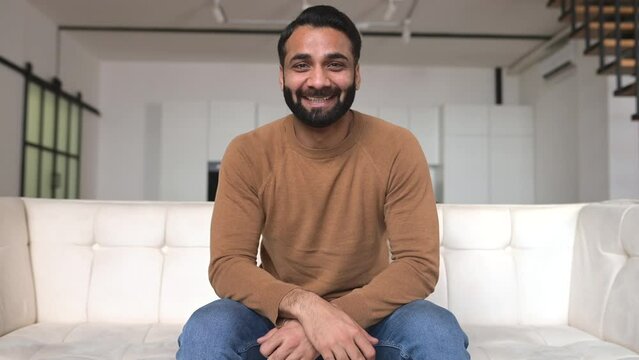 Smiling friendly young bearded Indian freelancer guy look and talks at camera, making video call to employee or friend, webcam view at ethnic male colleague involved virtual meeting sitting at home