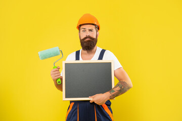 confused bearded man in helmet and work clothes hold paint roller and blackboard