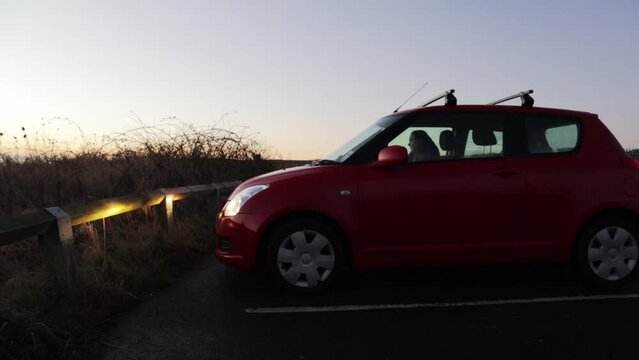 Small red car with headlights on pulling into a car parking space at dawn or dusk