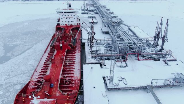 A large red ship tanker stands at the pier in the port and refuels in winter in the midst of snow and ice, aerial view