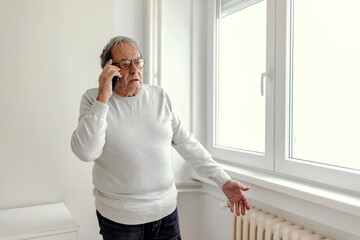 A man standing by a large window in the bedroom calls a repair service due to heating problems. An...