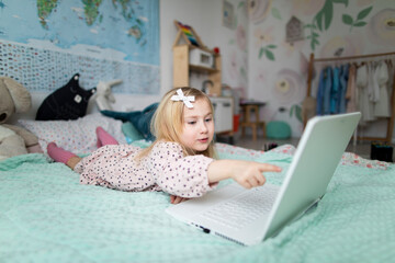 A little girl 5 years old lies on a bed in a children's room and looks into a white laptop and is surprised at what she sees there