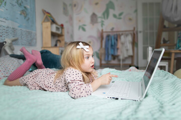 A little girl 5 years old lies on a bed in a children's room and looks into a white laptop and is surprised at what she sees there