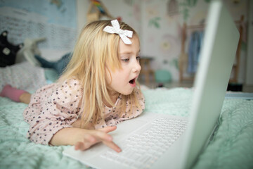 A little girl 5 years old lies on a bed in a children's room and looks into a white laptop and is surprised at what she sees there