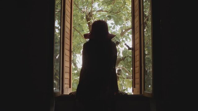 Young girl in a red hat with long curly hair opens a wooden window and leans on the windowsill. View from behind.