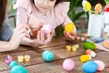 Happy Easter atmosphere! Close up funny, pretty, creative mom teacher, explain, to her cute, small, joyful daughter child how to draw, paint, decorate Easter eggs, together, sitting at desk
