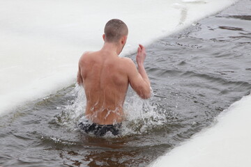 Young man bathes in icy water with splashes in an ice hole. Winter water sport.