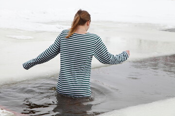 A slender European woman in a striped vest bathes in icy water in an ice hole in winter. Winter...