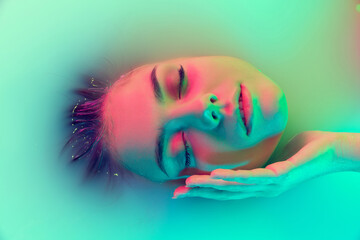 Top view of female face, young girl in the milk bath with soft glowing in blue-green neon light....