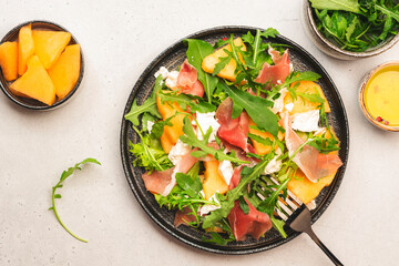 Summer melon salad with cantaloupe, prosciutto, soft cheese and arugula on white table background,...