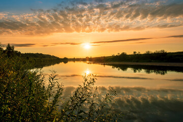 Naklejka na ściany i meble Scenic view at beautiful sunset or sunrise on a shiny river with green bushes on sides, golden sun rays, calm water ,deep blue cloudy sky and forest on a background, spring landscape