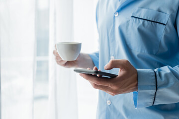 partial view of man in blue pajamas holding smartphone and coffee cup.