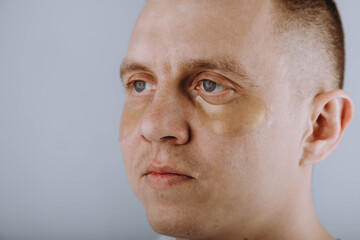 A serious man with hydrogel patches applied under his eyes looks at the camera. Individual morning skin care, anti-wrinkle, moisturizing cream, refreshing facial skin care concept 