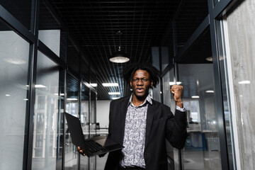 Happy black man clenched fists and rejoice finishing his online job on laptop. Working online in coworking space.