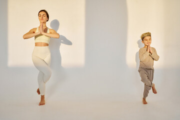 brunette woman with her blonde child son making yoga poses on white background, sunset light in studio