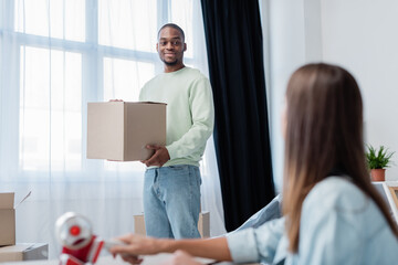 african american man holding box and looking at blurred woman at home.