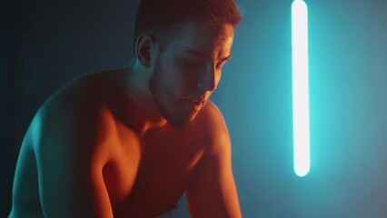 A tired athlete is resting in breathing heavily while sitting on a barbell in neon light after performing an exercise in slow motion. Recuperate and prepare for a new breakthrough