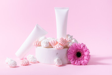 Creative composition with two white unbranded cosmetic tubes on the podium with bright gerbra daisy flower and merengue cookies on light pink background. Cosmetic branding banner concept.
