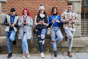 Full-body multiethnic men and women in casual clothing using cell phones while leaning against a...