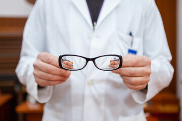 An eye doctor in a white coat shows a pair of corrective eyeglasses to correct myopia. The glasses have a black frame and the hands of the optician and the background are blurry.