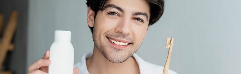 Fototapeta premium cheerful man holding toothpaste and toothbrush while looking at camera, banner.