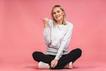 Portrait of a young beautiful cute cheerful blonde woman smiling isolated over pink background. Pointing fingers up. Sitting on the floor in a lotus pose.
