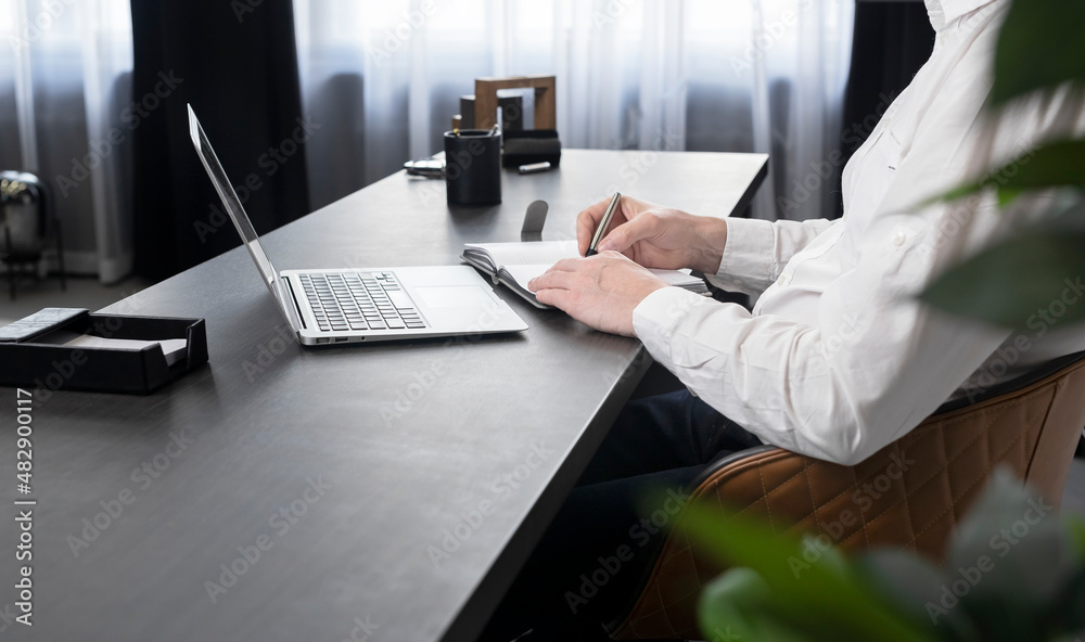 Poster man writing in notebook, using laptop at office desk.