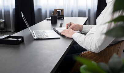 Man writing in notebook, using laptop at office desk.