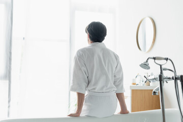 back view of brunette man in white bathrobe sitting on bathtub.