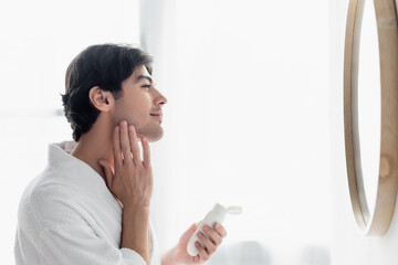 side view of brunette man applying cosmetic milk on face in bathroom.