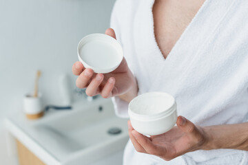 Fototapeta na wymiar cropped view of man in white bathrobe holding cosmetic cream in blurred bathroom.