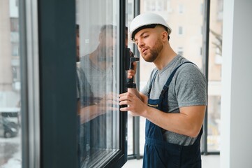 Workman in overalls installing or adjusting plastic windows in the living room at home