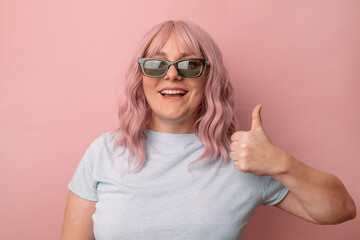 Happy young caucasian woman in bright stylish sunglasses and a blue t-shirt showing thumbs up and smiling isolated over pink background.