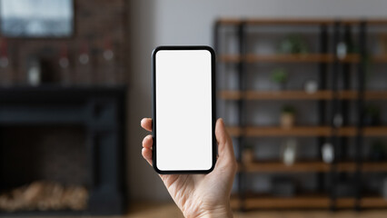 Female hand holds smartphone with white blank mock up screen, close up, living room on blurred background. Modern tech, new mobile application ad, on-line retail services, e-commerce usage concept