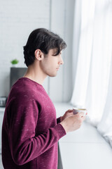 side view of young brunette man standing with coffee cup near window.