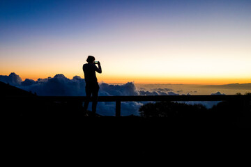Silhouette of a woman photographs a sunset on top of a mountain above the clouds