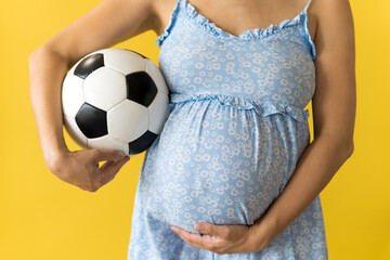 Motherhood, femininity, football, sport, dairy, hot summer. croped unrecognizable pregnant young pretty woman in floral blue dress holds soccer white and black ball rubs tummy on yellow background