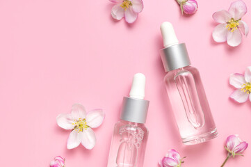 Clean glass cosmetic bottles with dropper, delicate spring flowers on pink background flat lay top view. Spring concept of natural organic cosmetics, beauty herbal product spa aroma oil. Mockup