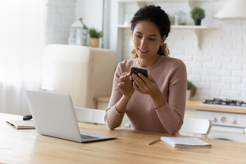Attractive 30s Latin woman sit at table with laptop in kitchen using smartphone. Make easy remote...