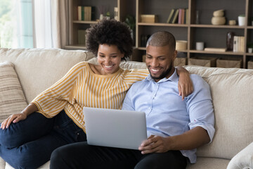 Beautiful cheerful African boyfriend and girlfriends relax on sofa with laptop, put wireless computer on laps watching streaming digital online TV, smiling enjoy video vlog, using e-services concept
