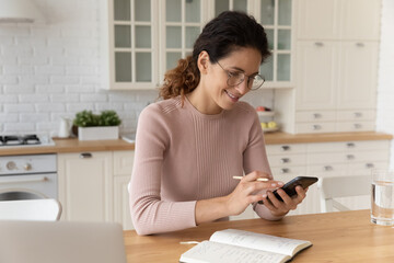 Young hispanic woman in eyeglasses sit at dining table holding smartphone and pencil, check...