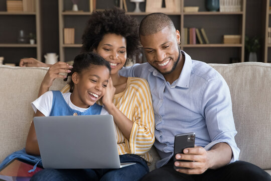 Young African couple and daughter sit on sofa watch videos on cell phone. 8s girl her mom and dad relaxing on couch with diverse electronic gadgets spend time on internet. Modern tech overuse concept