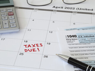 A 2022 calendar noting the April 18 USA Internal Revenue Service IRS income filing deadline for year 2021 taxes is shown up close, with a calculator, ink pen, eye glasses, and Form 1040 paperwork.