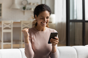 Excited 30s Latina woman sit on sofa with smartphone read great news feels happy look overjoyed got...
