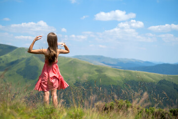 Young happy woman traveler in red dress standing on green grassy hillside on a windy day in summer mountains enjoying view of nature