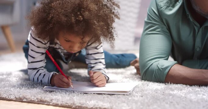 Close up of little beautiful african american girl with curly hair lying on the floor and carefully drawing picture in album.