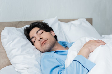 Fototapeta na wymiar brunette man in blue pajamas sleeping on white bedding in morning.