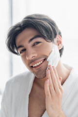 cheerful brunette man smiling at camera while applying shaving foam.