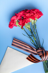 a bouquet of red carnations with a St. George ribbon and a triangular military envelope a field mail symbol with space for text on a blue background, top view, holiday background, postcard Defender of